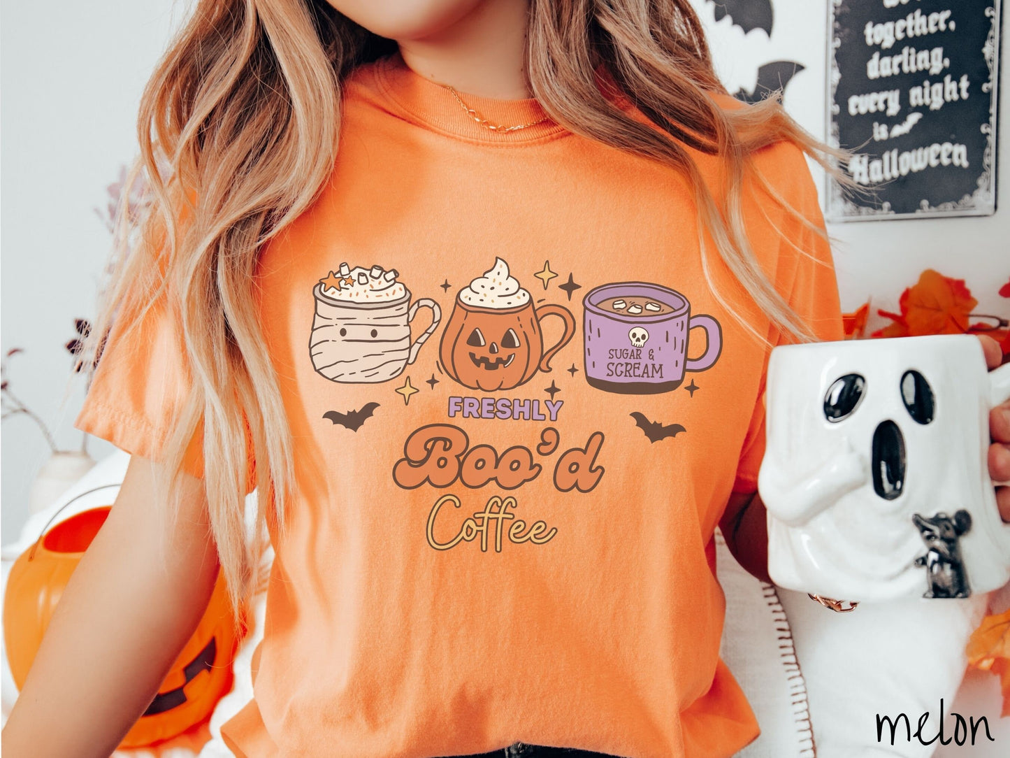 A woman wearing a cute, vintage melon colored Comfort Colors T-shirt with the text Freshly Boo’d Coffee. Above are coffee mugs where one looks like a mummy with whipped cream, one a carved pumpkin mug, and the last is purple with a skull.