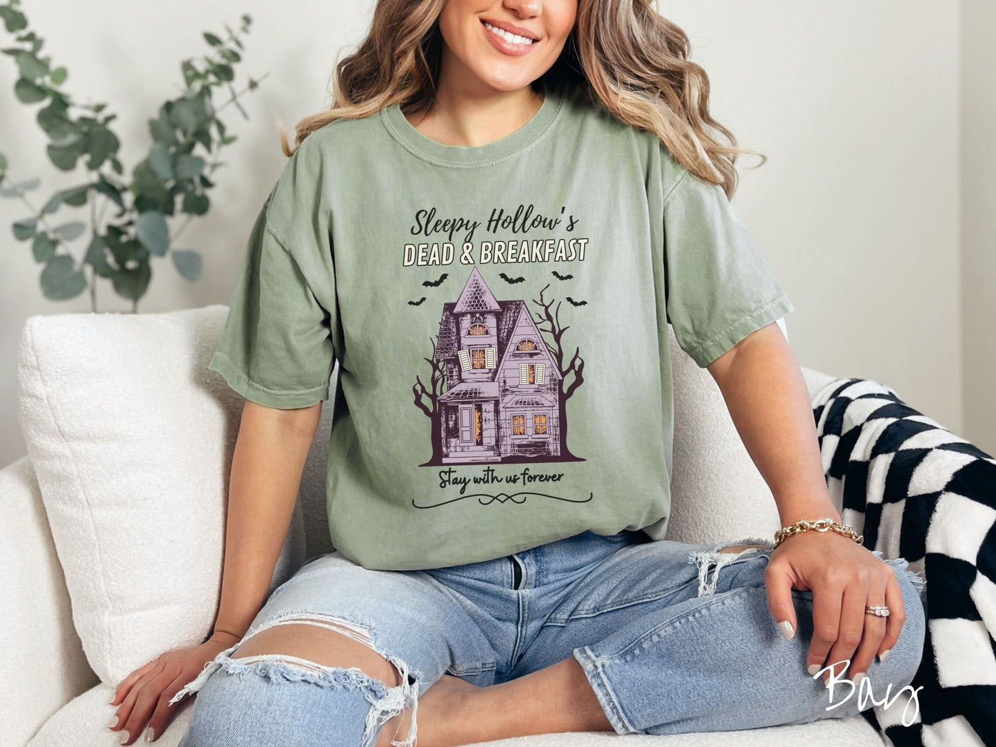 A woman wearing a cute, vintage bay colored Comfort Colors T-shirt with the text Sleepy Hollow’s Dead & Breakfast above then Stay with us forever below. In between the text is a spooky haunted house surrounded by black bats.