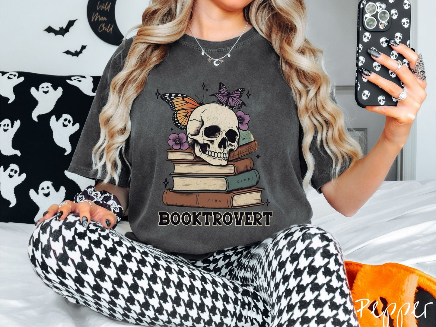 A woman wearing a cute, vintage pepper colored Comfort Colors T-shirt with the text Booktrovert in black font. Above are a stack of brown and green old books with a smiling skeleton skull sitting on top with colorful butterflies and flowers.