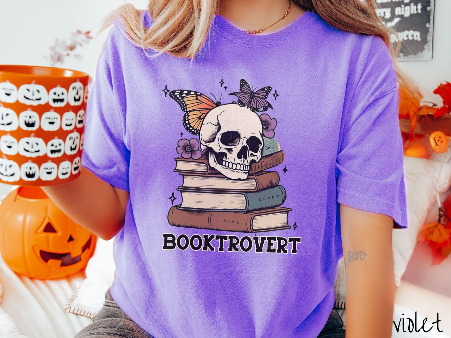 A woman wearing a cute, vintage violet colored Comfort Colors T-shirt with the text Booktrovert in black font. Above are a stack of brown and green old books with a smiling skeleton skull sitting on top with colorful butterflies and flowers.