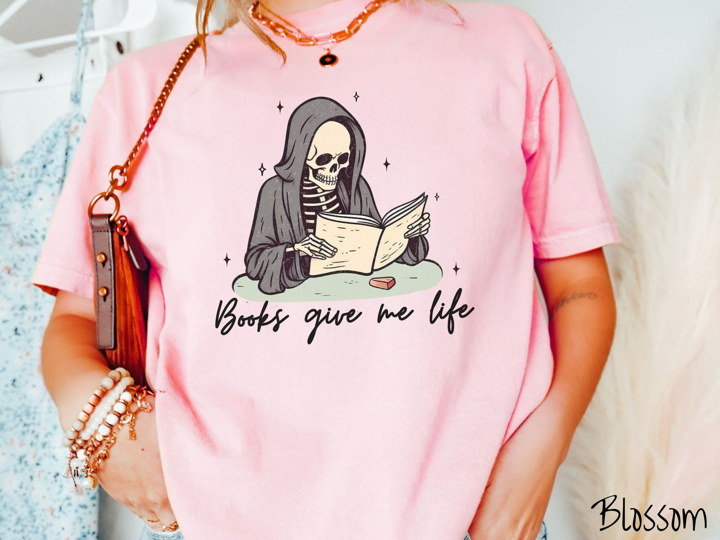 A woman wearing a cute, vintage blossom colored Comfort Colors T-shirt with the text Books give me life in black cursive font. Above is a smiling skeleton wearing a black hood reading a book surrounded by twinkling stars.