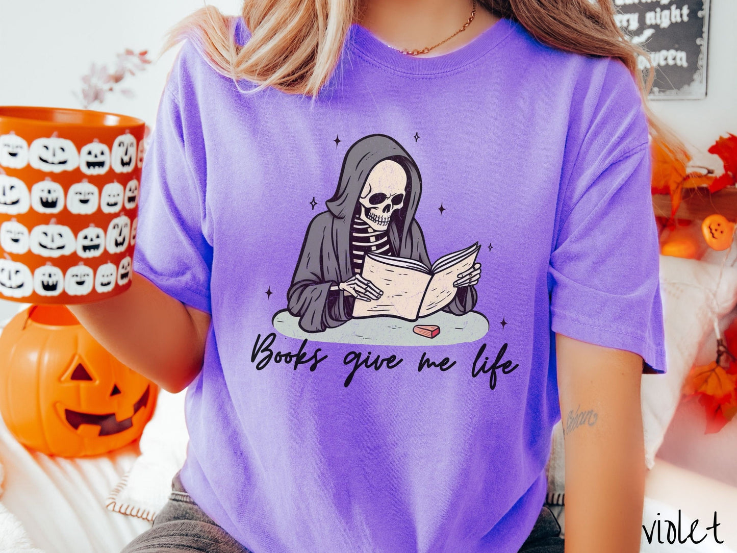 A woman wearing a cute, vintage violet colored Comfort Colors T-shirt with the text Books give me life in black cursive font. Above is a smiling skeleton wearing a black hood reading a book surrounded by twinkling stars.