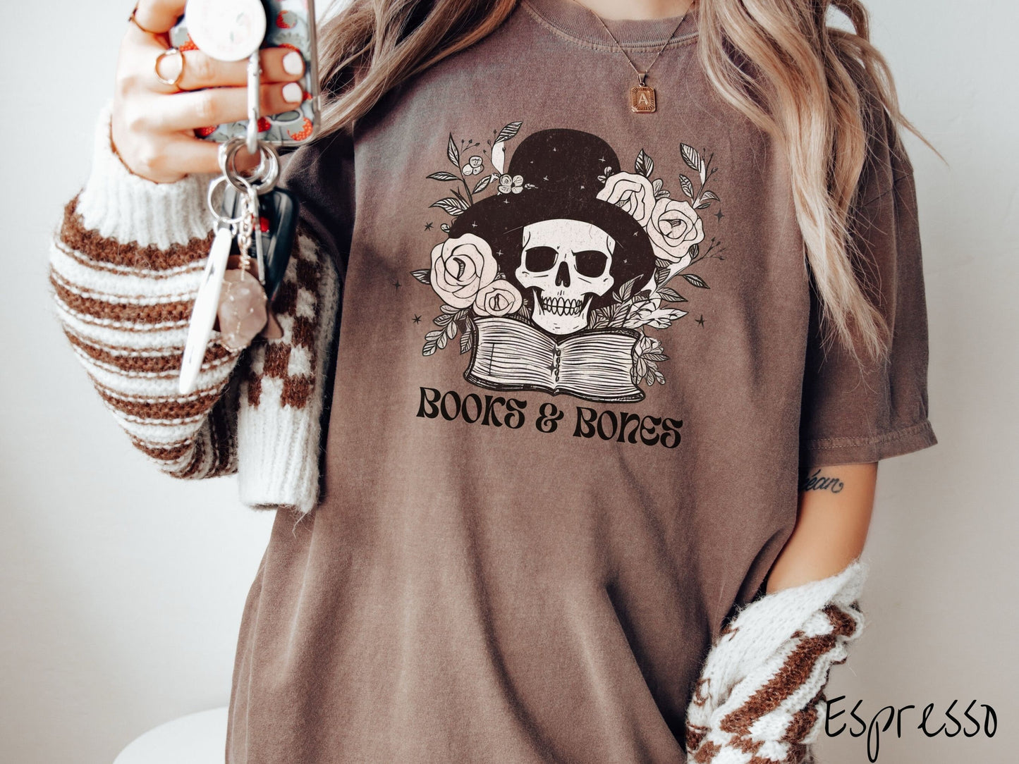 A woman wearing a cute, vintage espresso colored Comfort Colors T-shirt with the text Books & Bones in black font. Above is a smiling skeleton wearing a black top hat surrounded by roses and leaves, and hovering above a thick, leather bound book.