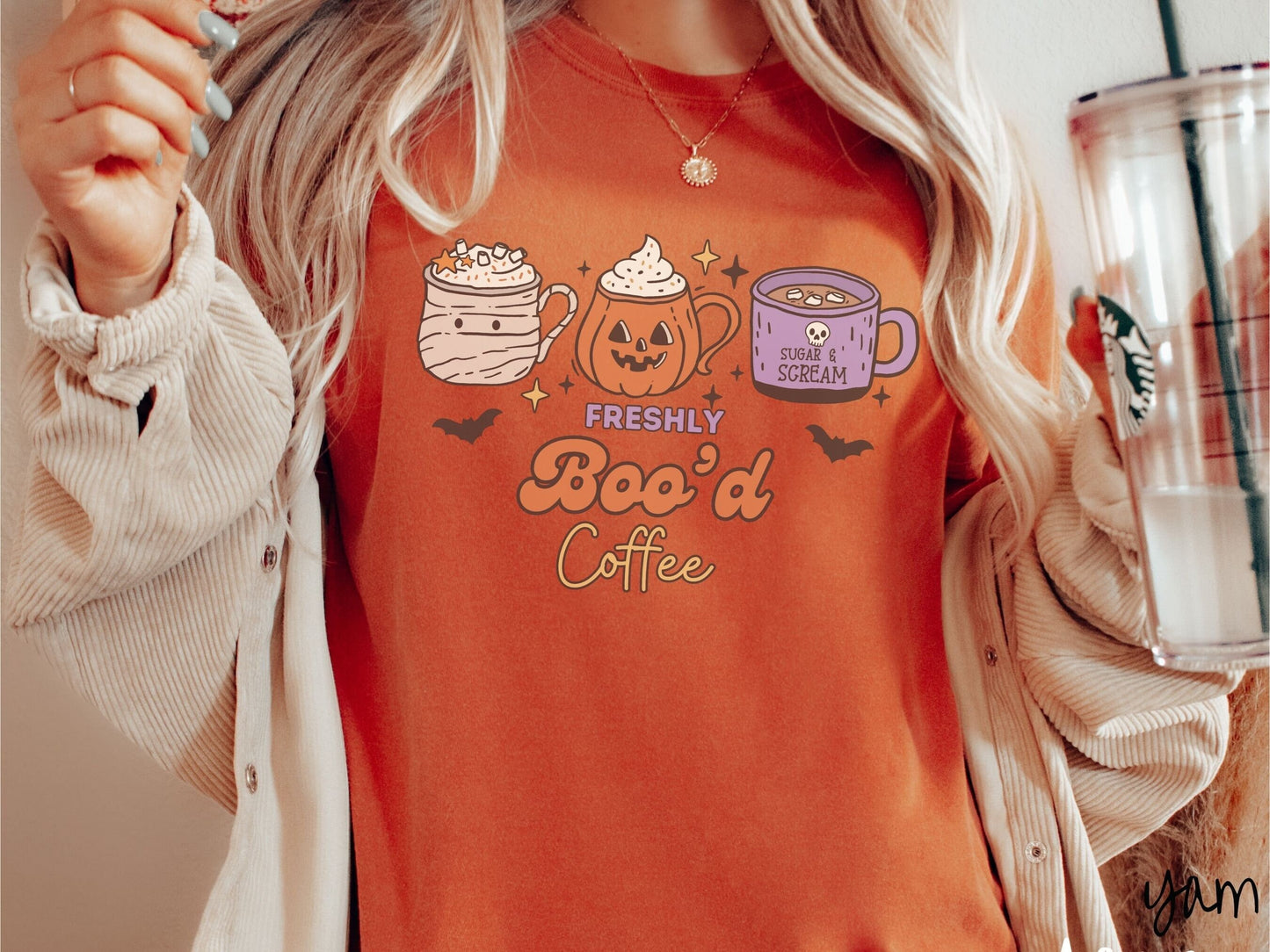 A woman wearing a cute, vintage yam colored Comfort Colors T-shirt with the text Freshly Boo’d Coffee. Above are coffee mugs where one looks like a mummy with whipped cream, one a carved pumpkin mug, and the last is purple with a skull.