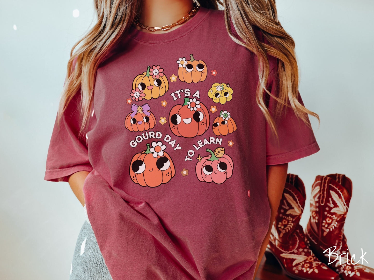 A woman wearing a cute, vintage brick colored Comfort Colors T-shirt with the text Its a Gourd Day to Learn in white font. There is a three by three grid of orange, yellow, and pink pumpkins with girly eyes, wearing flowers and bows.