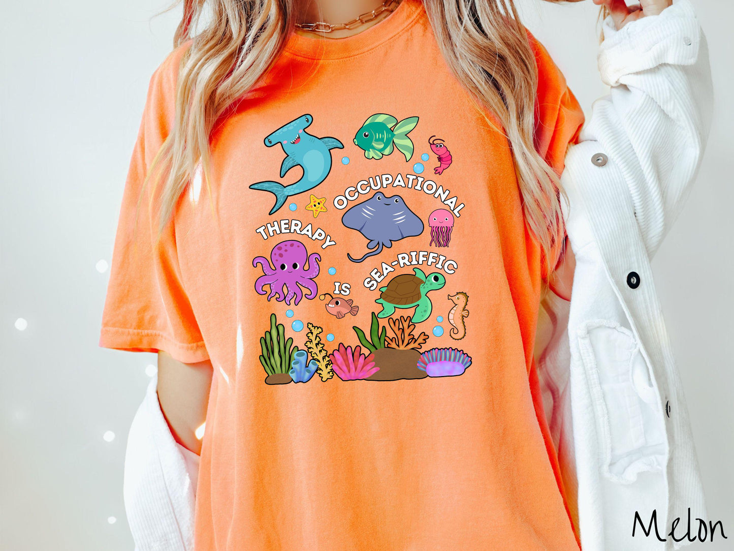 A woman wearing a cute, vintage melon colored Comfort Colors T-shirt with the text Occupational Therapy is Sea-Rific. There is a shark, green fish, octopus, stingray, jellyfish, turtle, and seahorse swimming together above a colorful coral reef.