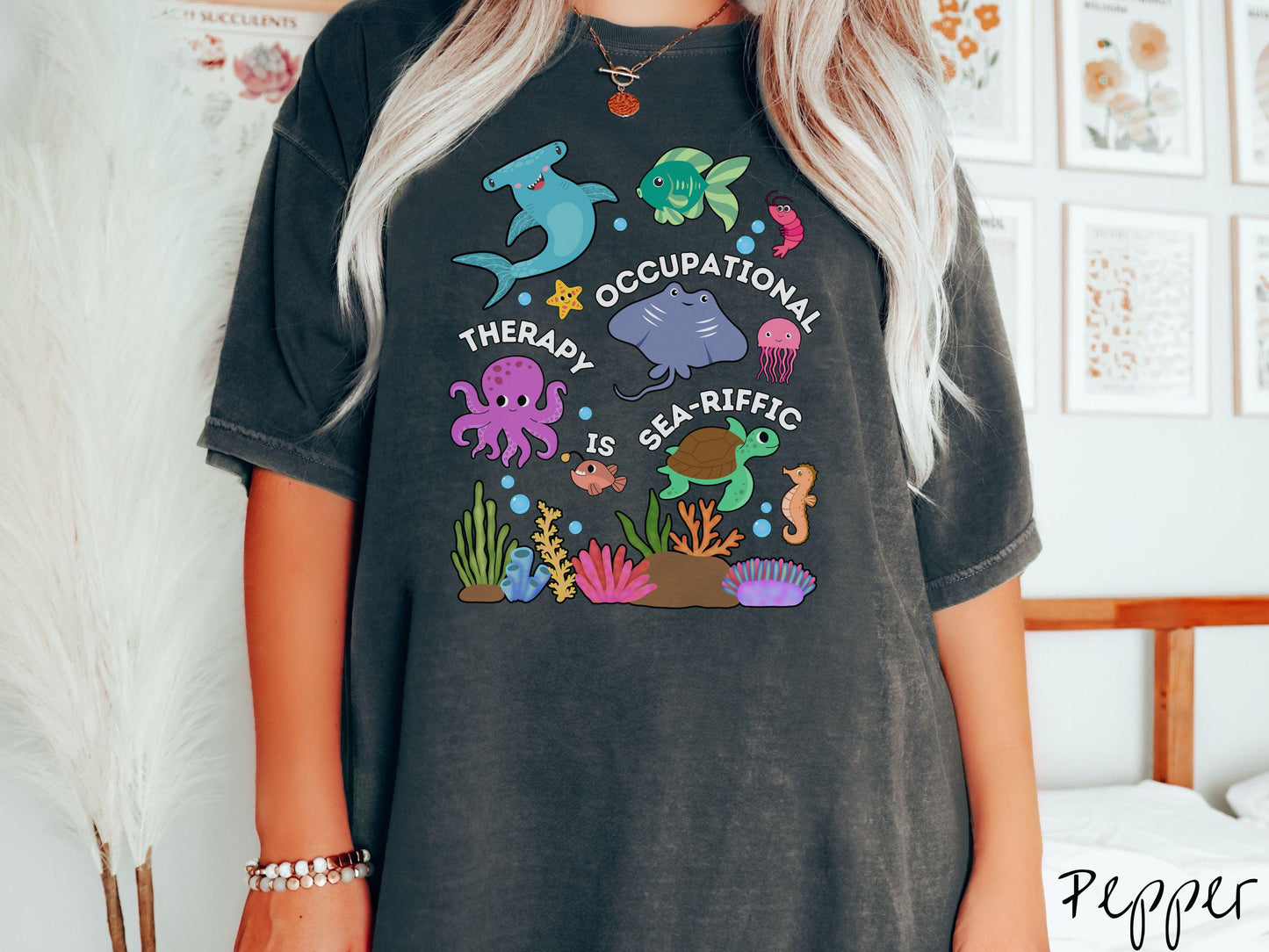 A woman wearing a cute, vintage pepper colored Comfort Colors T-shirt with the text Occupational Therapy is Sea-Rific. There is a shark, green fish, octopus, stingray, jellyfish, turtle, and seahorse swimming together above a colorful coral reef.
