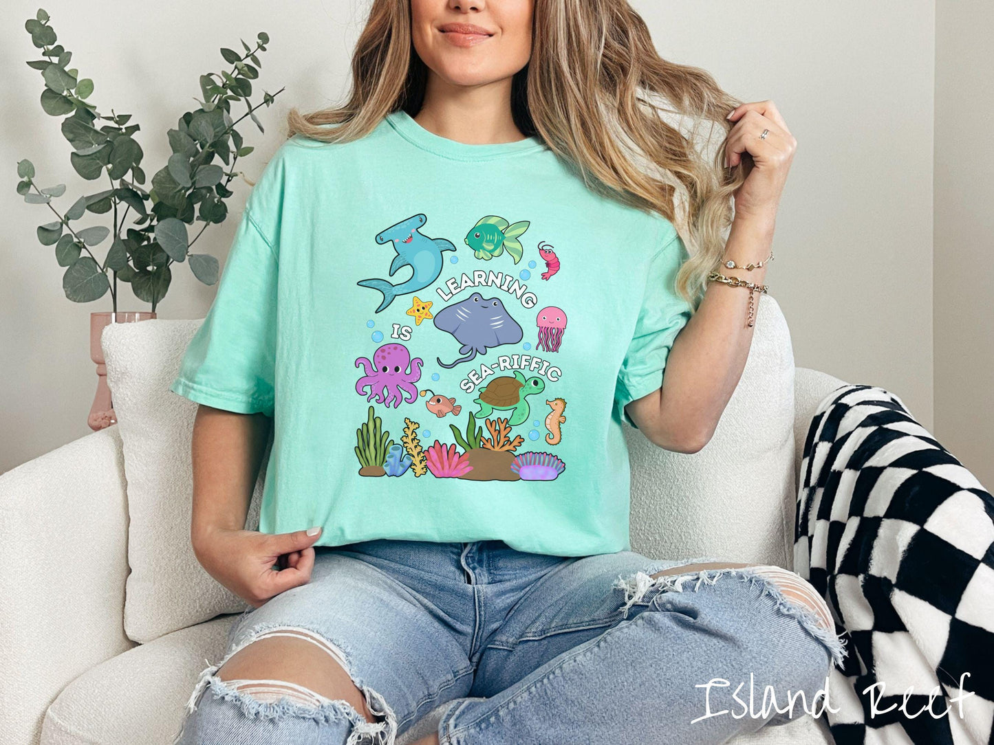 A woman wearing a cute, vintage island reef colored Comfort Colors T-shirt with the text Learning is Sea-Rific. There is a shark, green fish, octopus, stingray, jellyfish, turtle, and seahorse swimming together above a colorful coral reef.