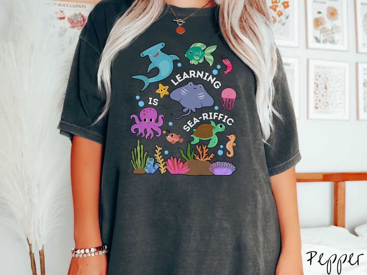 A woman wearing a cute, vintage pepper colored Comfort Colors T-shirt with the text Learning is Sea-Rific. There is a shark, green fish, octopus, stingray, jellyfish, turtle, and seahorse swimming together above a colorful coral reef.