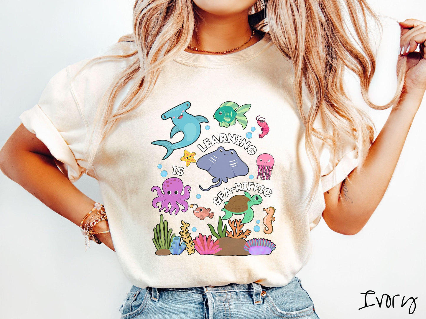 A woman wearing a cute, vintage ivory colored Comfort Colors T-shirt with the text Learning is Sea-Rific. There is a shark, green fish, octopus, stingray, jellyfish, turtle, and seahorse swimming together above a colorful coral reef.