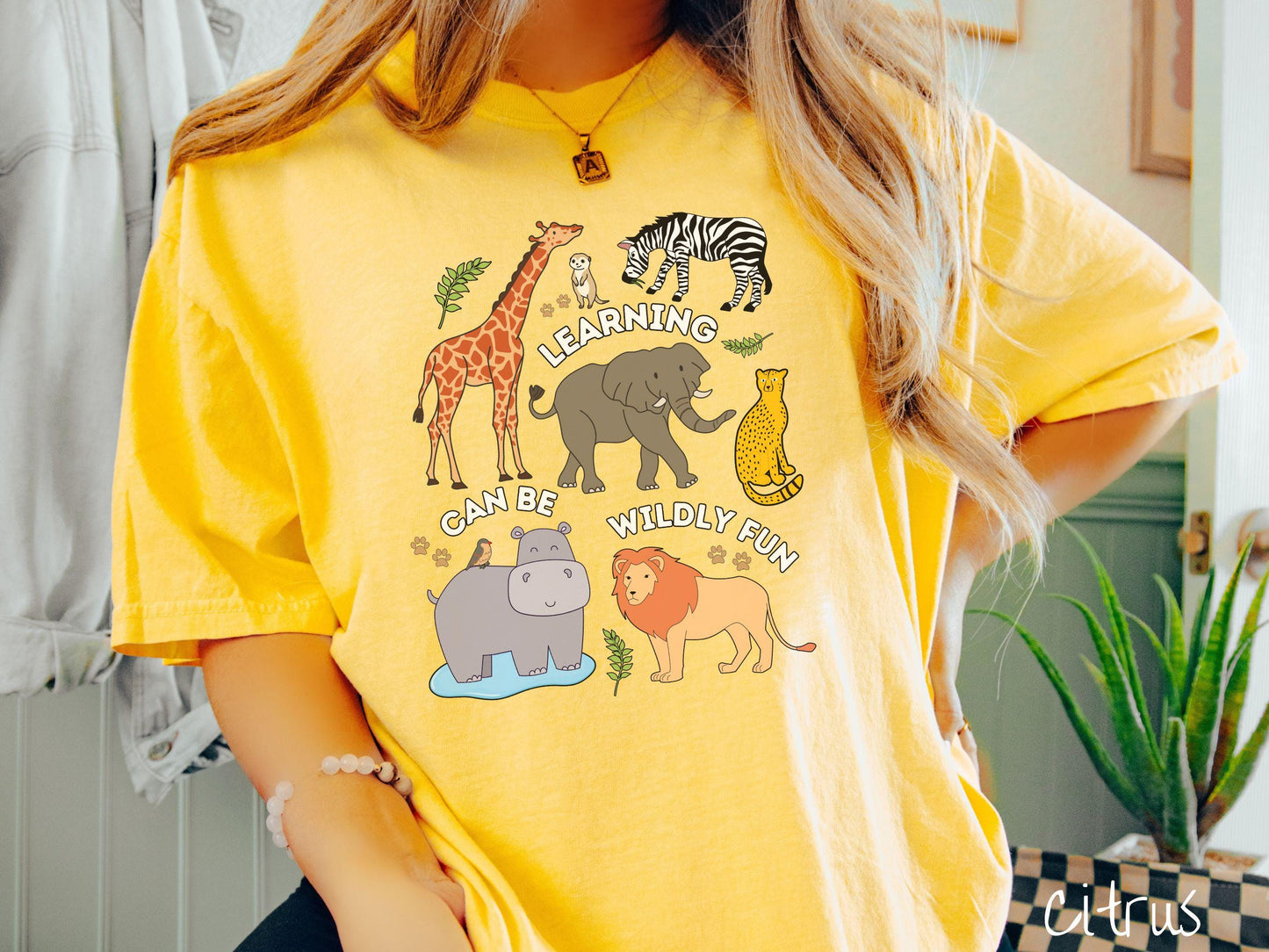 A woman wearing a cute, vintage citrus colored Comfort Colors T-shirt with the text Learning Can Be Wildly Fun. There is a giraffe, zebra elephant, hippopotamus, lion, leopard, and ground squirrel surrounded by leafy branches and paw prints.