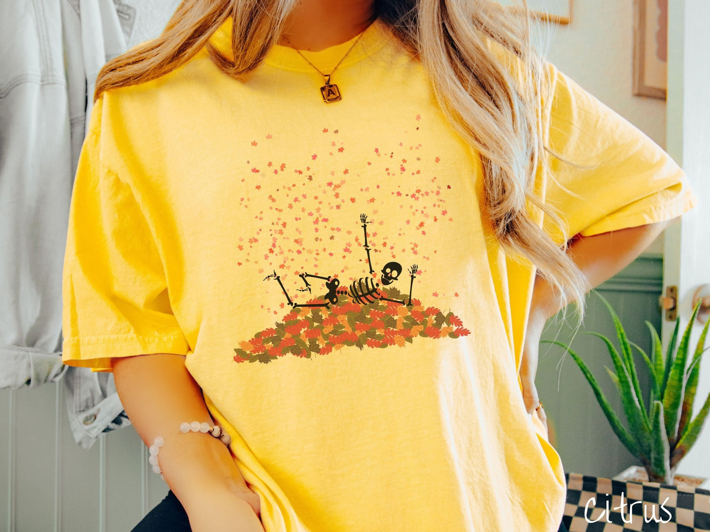 A woman wearing a cute, vintage citrus colored Comfort Colors T-shirt with a black skeleton playing in a pile of colorful fall leaves. The skeleton is throwing leaves in the air, and there are colorful leaves floating above.