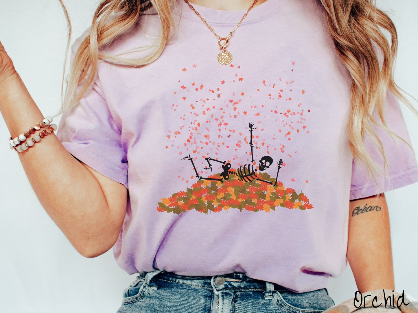 A woman wearing a cute, vintage orchid colored Comfort Colors T-shirt with a black skeleton playing in a pile of colorful fall leaves. The skeleton is throwing leaves in the air, and there are colorful leaves floating above.