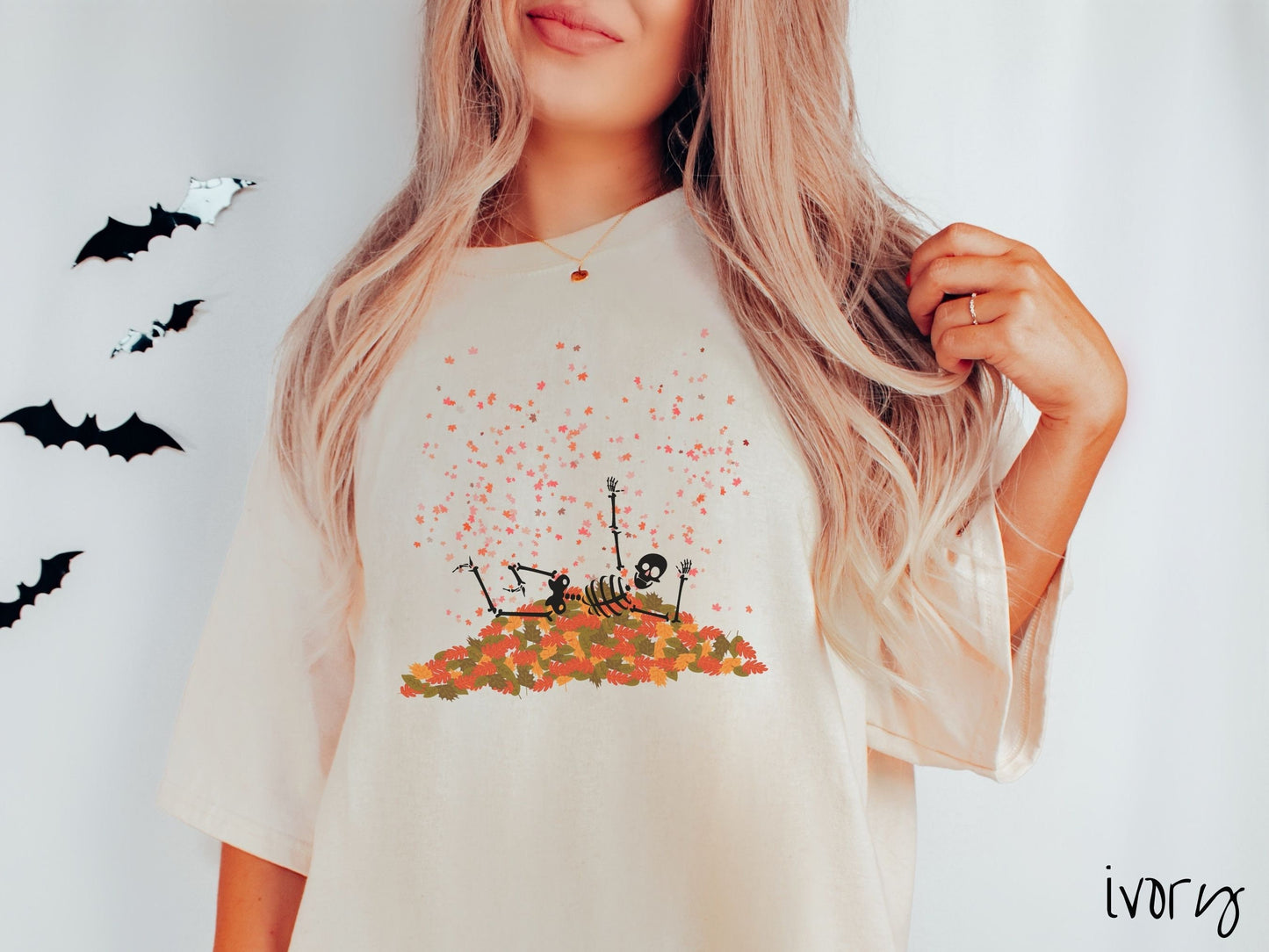 A woman wearing a cute, vintage ivory colored Comfort Colors T-shirt with a black skeleton playing in a pile of colorful fall leaves. The skeleton is throwing leaves in the air, and there are colorful leaves floating above.