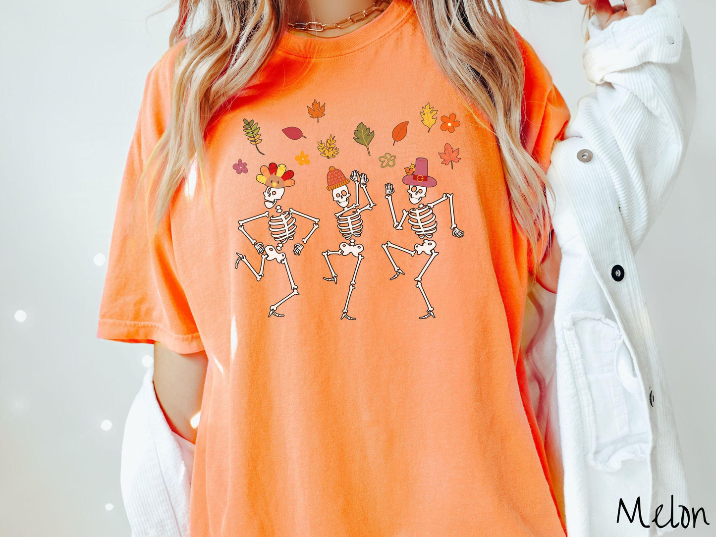 A woman wearing a cute melon colored Comfort Colors t-shirt with three white skeletons wearing pilgrim hats dancing with colorful fall leaves in the air.