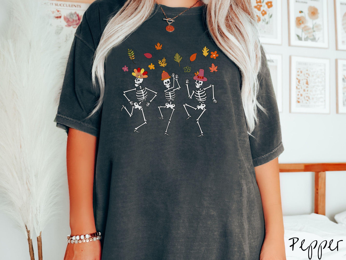 A woman wearing a cute pepper colored Comfort Colors t-shirt with three white skeletons wearing pilgrim hats dancing with colorful fall leaves in the air.