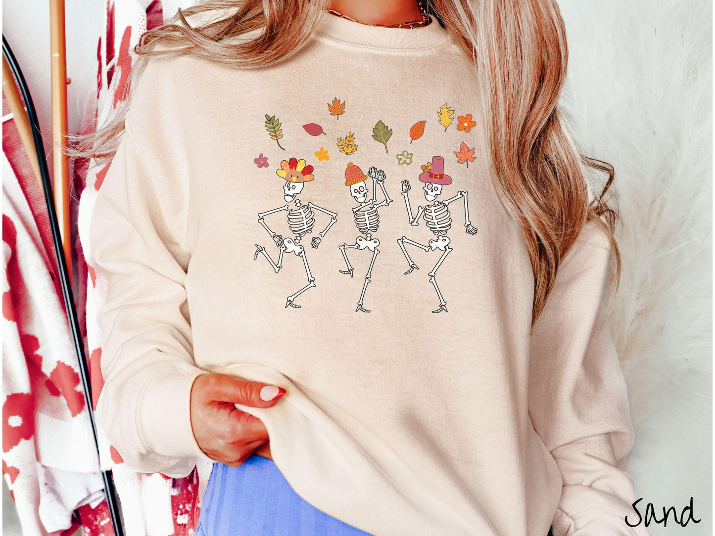 A woman wearing a cute sand colored sweatshirt with three white skeletons wearing pilgrim hats dancing with colorful fall leaves in the air.