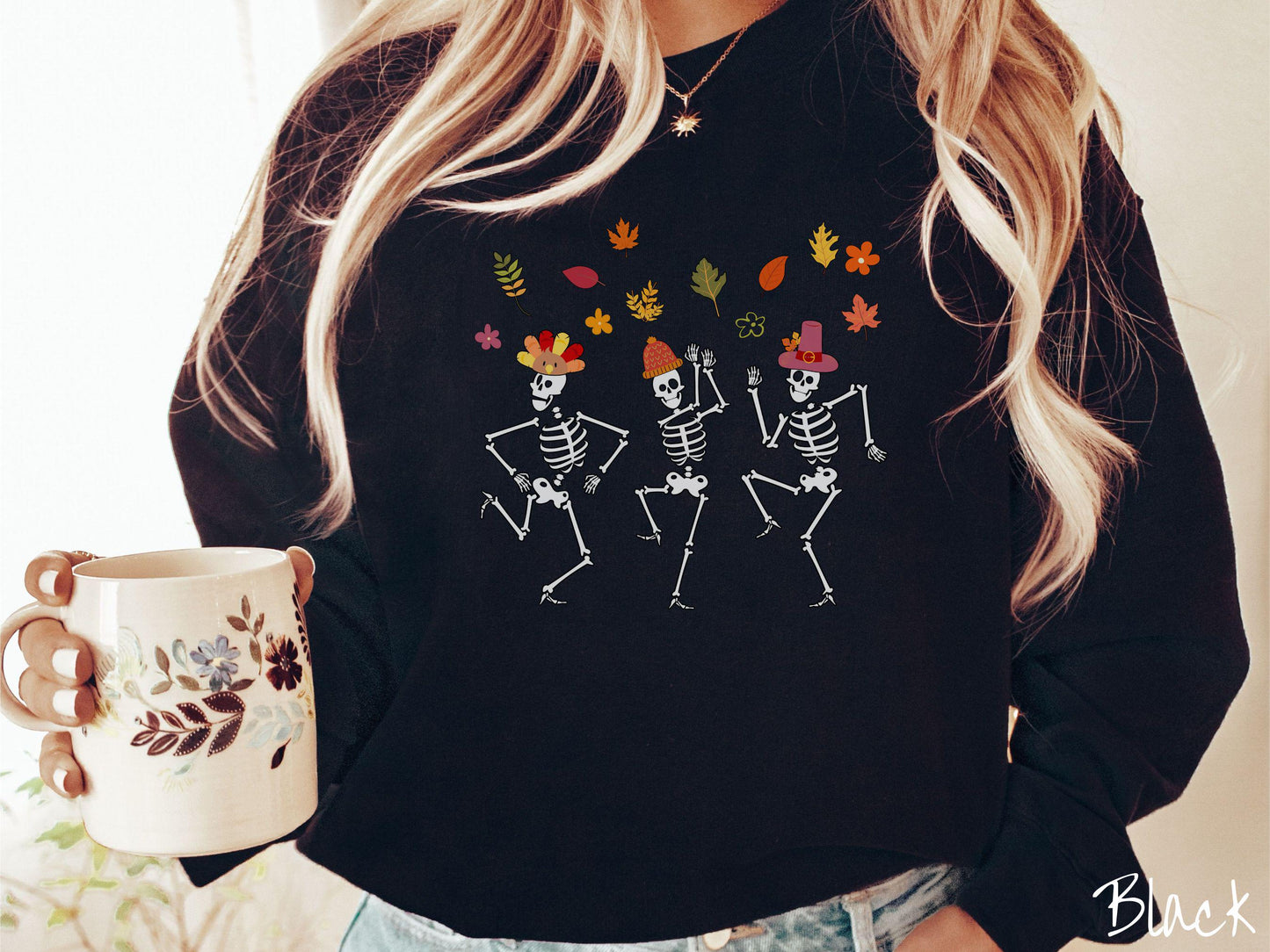 A woman wearing a cute black colored sweatshirt with three white skeletons wearing pilgrim hats dancing with colorful fall leaves in the air.