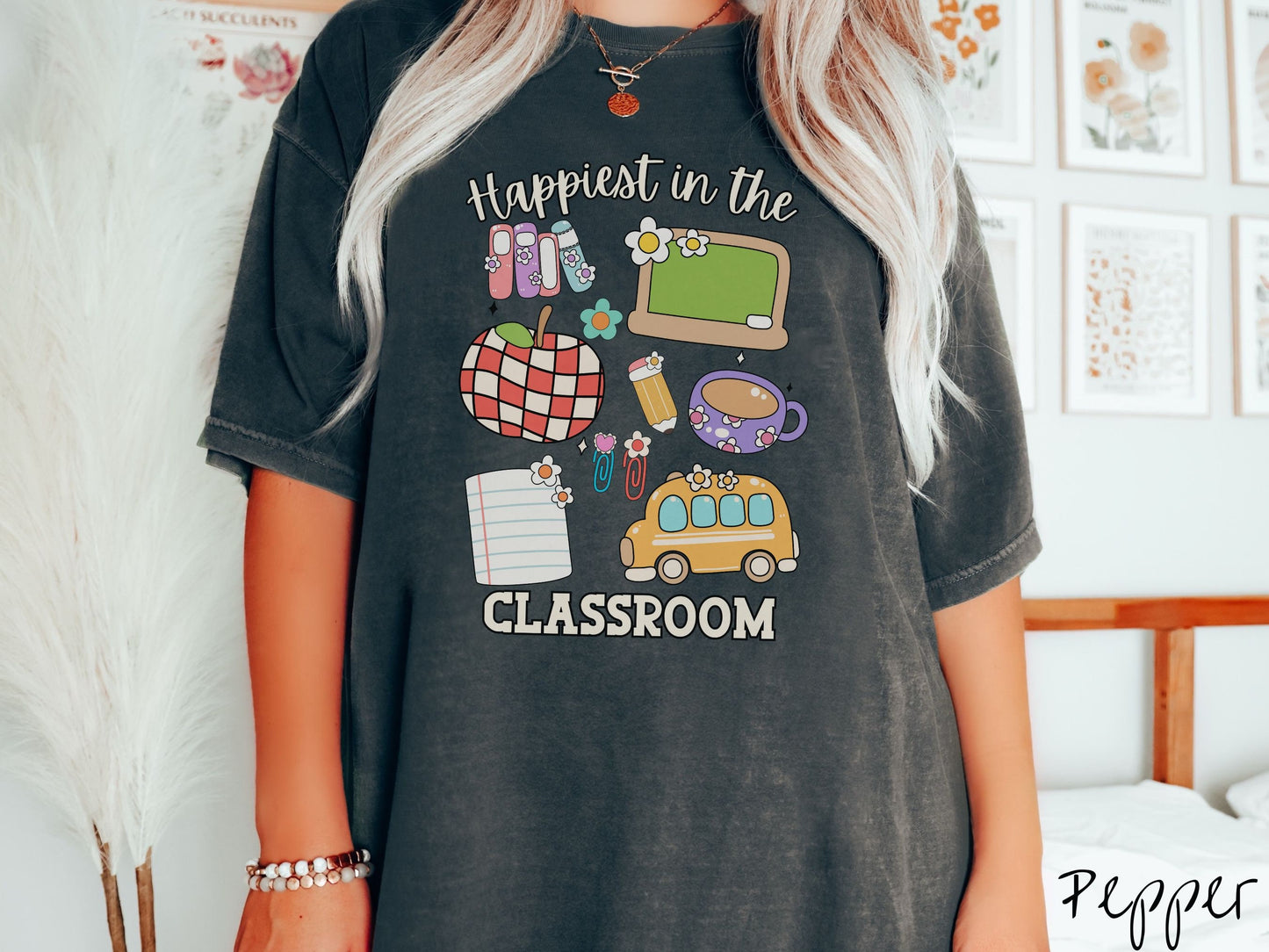 A woman wearing a cute, vintage pepper colored Comfort Colors T-shirt with the text Happiest in the Classroom. In between the text are books, an apple, chalkboard, pencil and paper, coffee cup, yellow school bus, paperclips, and flowers.