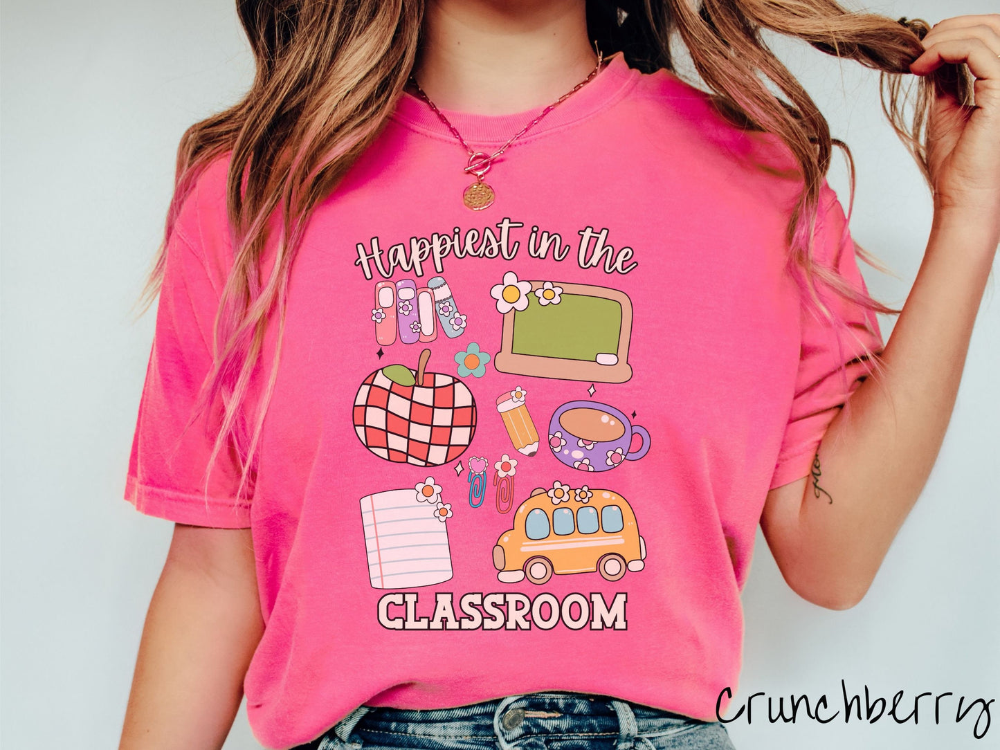 A woman wearing a cute, vintage crunchberry colored Comfort Colors T-shirt with the text Happiest in the Classroom. In between the text are books, an apple, chalkboard, pencil and paper, coffee cup, yellow school bus, paperclips, and flowers.