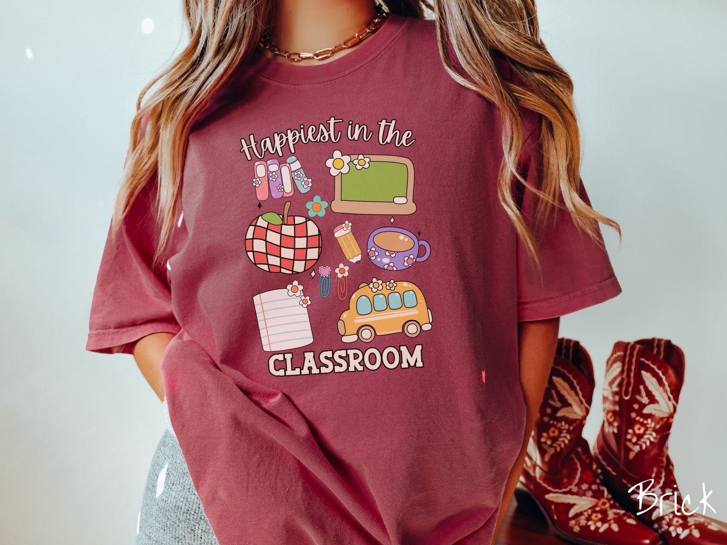 A woman wearing a cute, vintage brick colored Comfort Colors T-shirt with the text Happiest in the Classroom. In between the text are books, an apple, chalkboard, pencil and paper, coffee cup, yellow school bus, paperclips, and flowers.