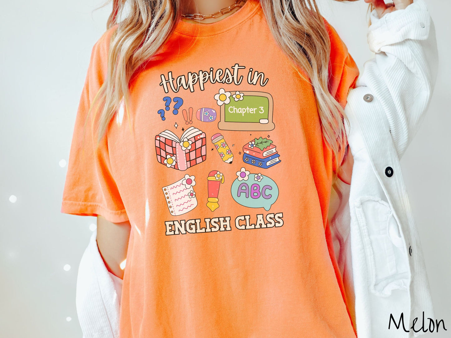 A woman wearing a cute, vintage melon colored Comfort Colors T-shirt with the text Happiest in English Class. In between the text are books, erasers, a chalkboard, pencil, pen, notebook paper, alphabet letters, and flowers.