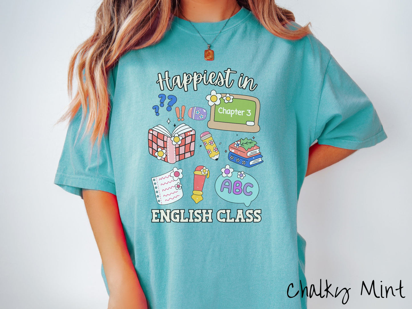A woman wearing a cute, vintage chalky mint colored Comfort Colors T-shirt with the text Happiest in English Class. In between the text are books, erasers, a chalkboard, pencil, pen, notebook paper, alphabet letters, and flowers.