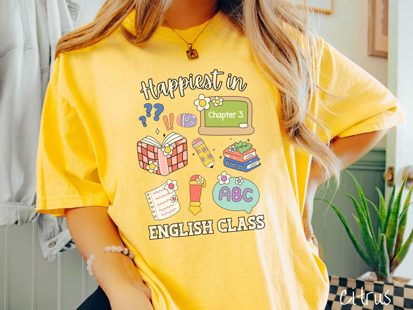 A woman wearing a cute, vintage citrus colored Comfort Colors T-shirt with the text Happiest in English Class. In between the text are books, erasers, a chalkboard, pencil, pen, notebook paper, alphabet letters, and flowers.