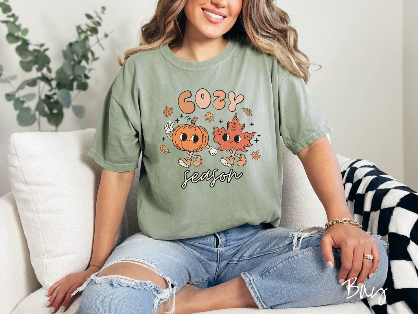 A woman wearing a cute, vintage bay colored Comfort Colors T-shirt with the text Cozy Season and in between the text are a smiling orange pumpkin holding up a peace sign with one hand and holding hands with a smiling dark orange leaf.