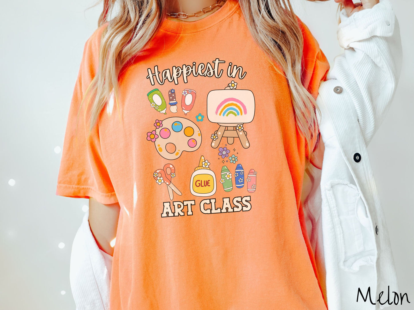 A woman wearing a cute, vintage melon colored Comfort Colors T-shirt with the text Happiest in Art Class. In between the text are paint brushes, a paint palette, an art canvas on a stand, scissors, glue, crayons, and flowers.