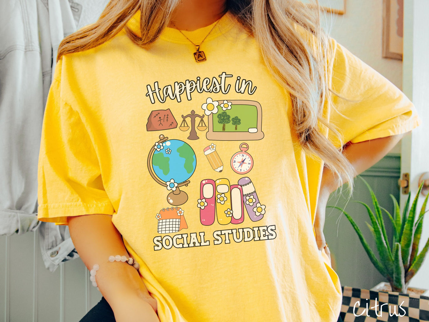 A woman wearing a cute, vintage citrus colored Comfort Colors T-shirt with the text Happiest in Social Studies Class. In between the text are hieroglyphics, a globe, a pencil, compass, books, laptop computer, and a weight scale.