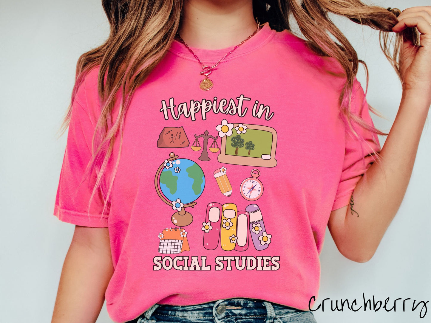 A woman wearing a cute, vintage crunchberry colored Comfort Colors T-shirt with the text Happiest in Social Studies Class. In between the text are hieroglyphics, a globe, a pencil, compass, books, laptop computer, and a weight scale.