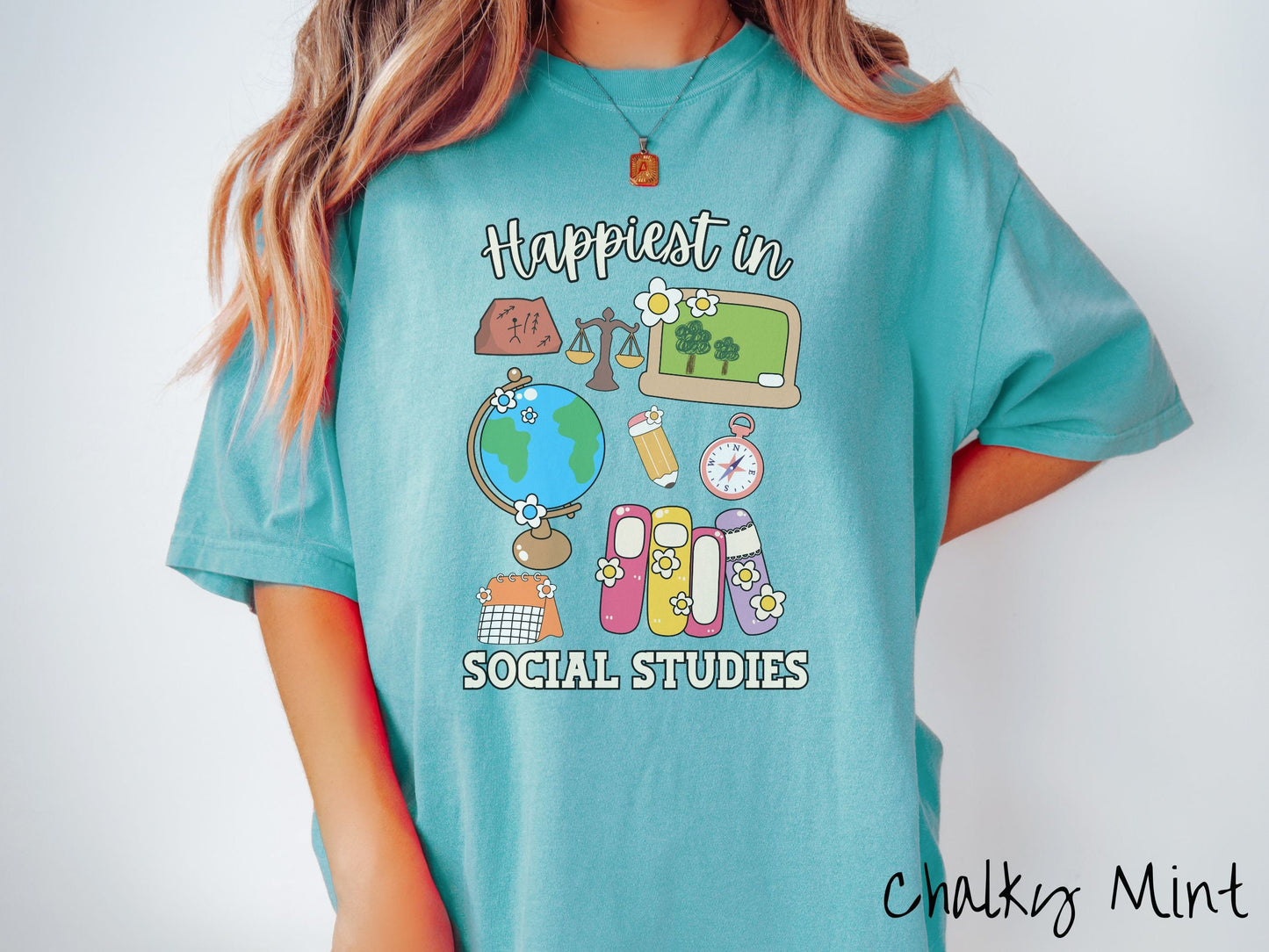 A woman wearing a cute, vintage chalky mint colored Comfort Colors T-shirt with the text Happiest in Social Studies Class. In between the text are hieroglyphics, a globe, a pencil, compass, books, laptop computer, and a weight scale.