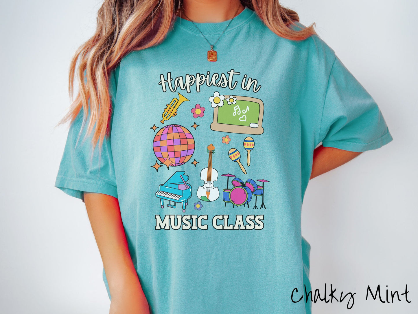 A woman wearing a cute, vintage chalky mint colored Comfort Colors T-shirt with the text Happiest in Music Class. In between the text are a saxophone, a disco ball, a chalkboard, a piano, a guitar, a drum set, maracas, and flowers.