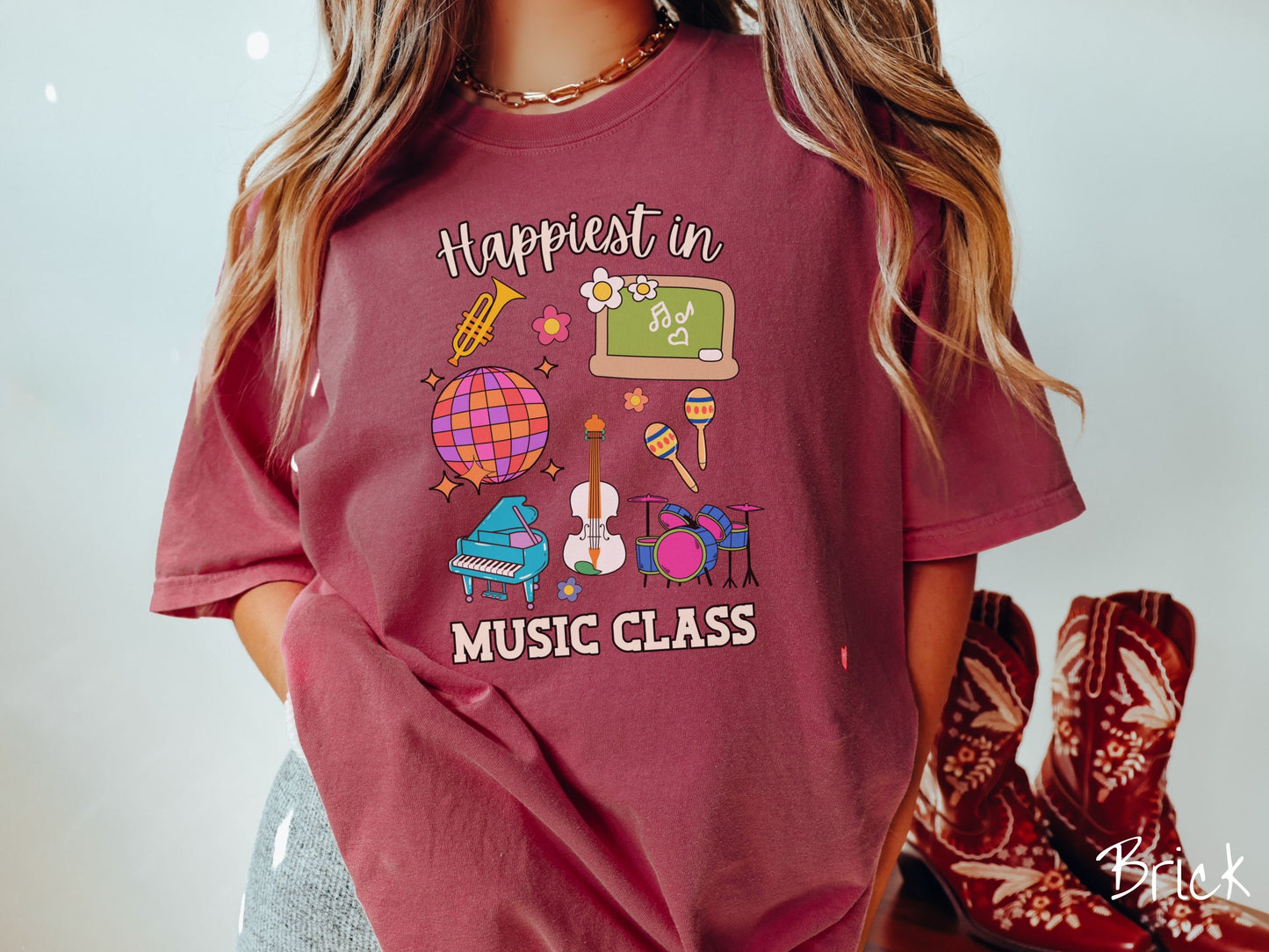 A woman wearing a cute, vintage brick colored Comfort Colors T-shirt with the text Happiest in Music Class. In between the text are a saxophone, a disco ball, a chalkboard, a piano, a guitar, a drum set, maracas, and flowers.
