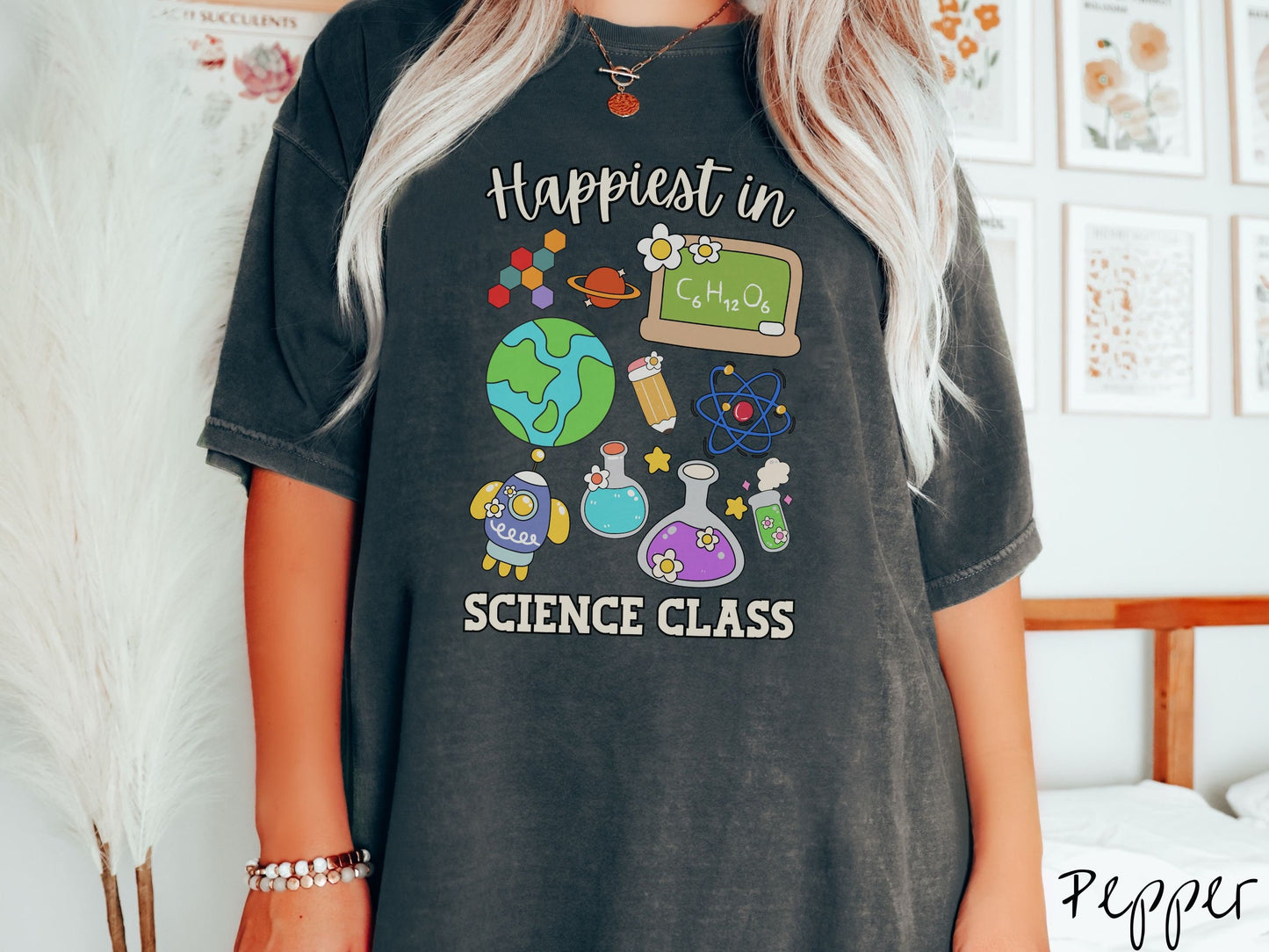 A woman wearing a cute, vintage pepper colored Comfort Colors T-shirt with the text Happiest in Science Class. In between the text are a globe, chemistry, rocket ship, chalkboard, science beakers and test tubes, a pencil, DNA molecules, and Saturn.