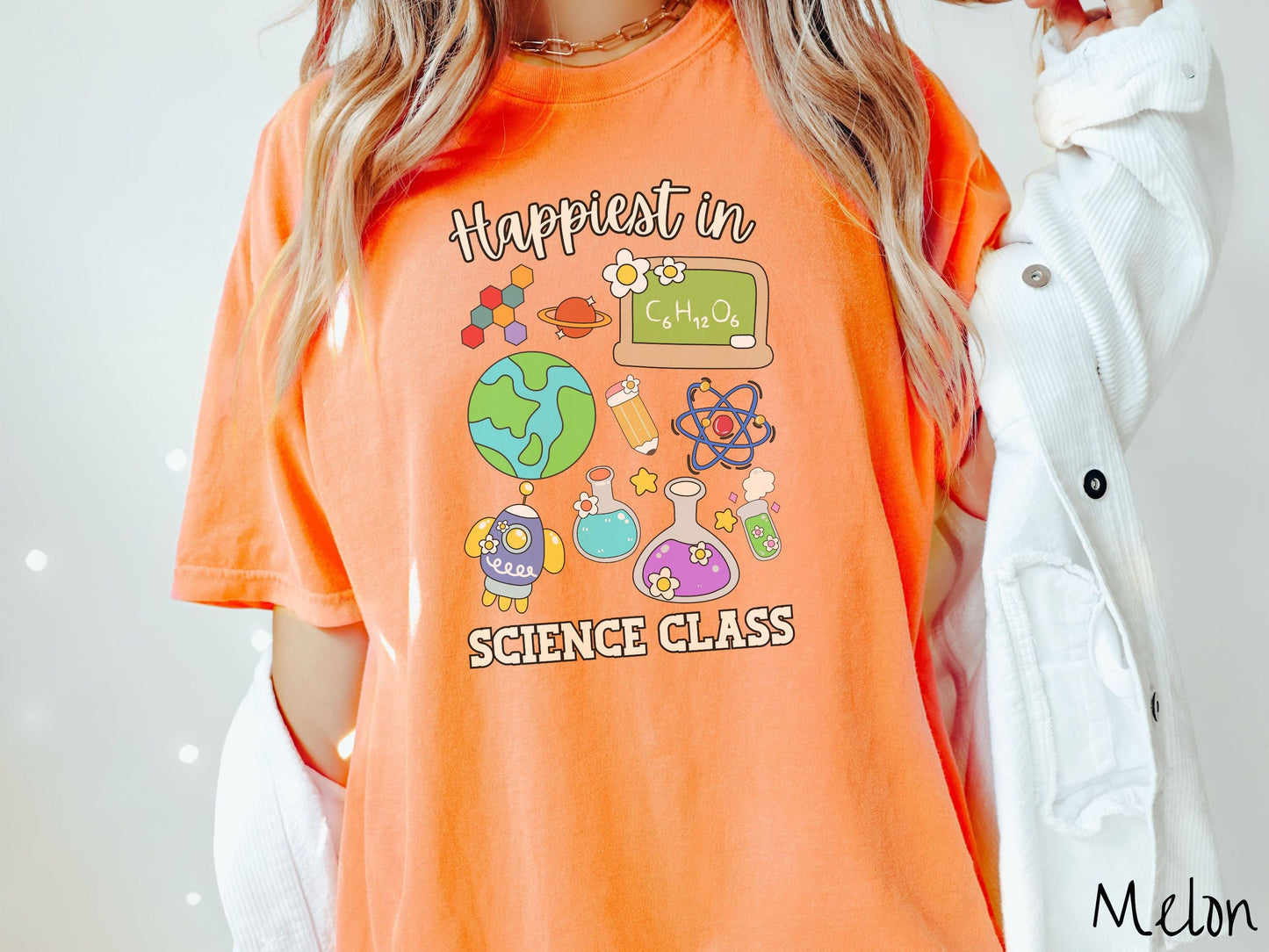 A woman wearing a cute, vintage melon colored Comfort Colors T-shirt with the text Happiest in Science Class. In between the text are a globe, chemistry, rocket ship, chalkboard, science beakers and test tubes, a pencil, DNA molecules, and Saturn.
