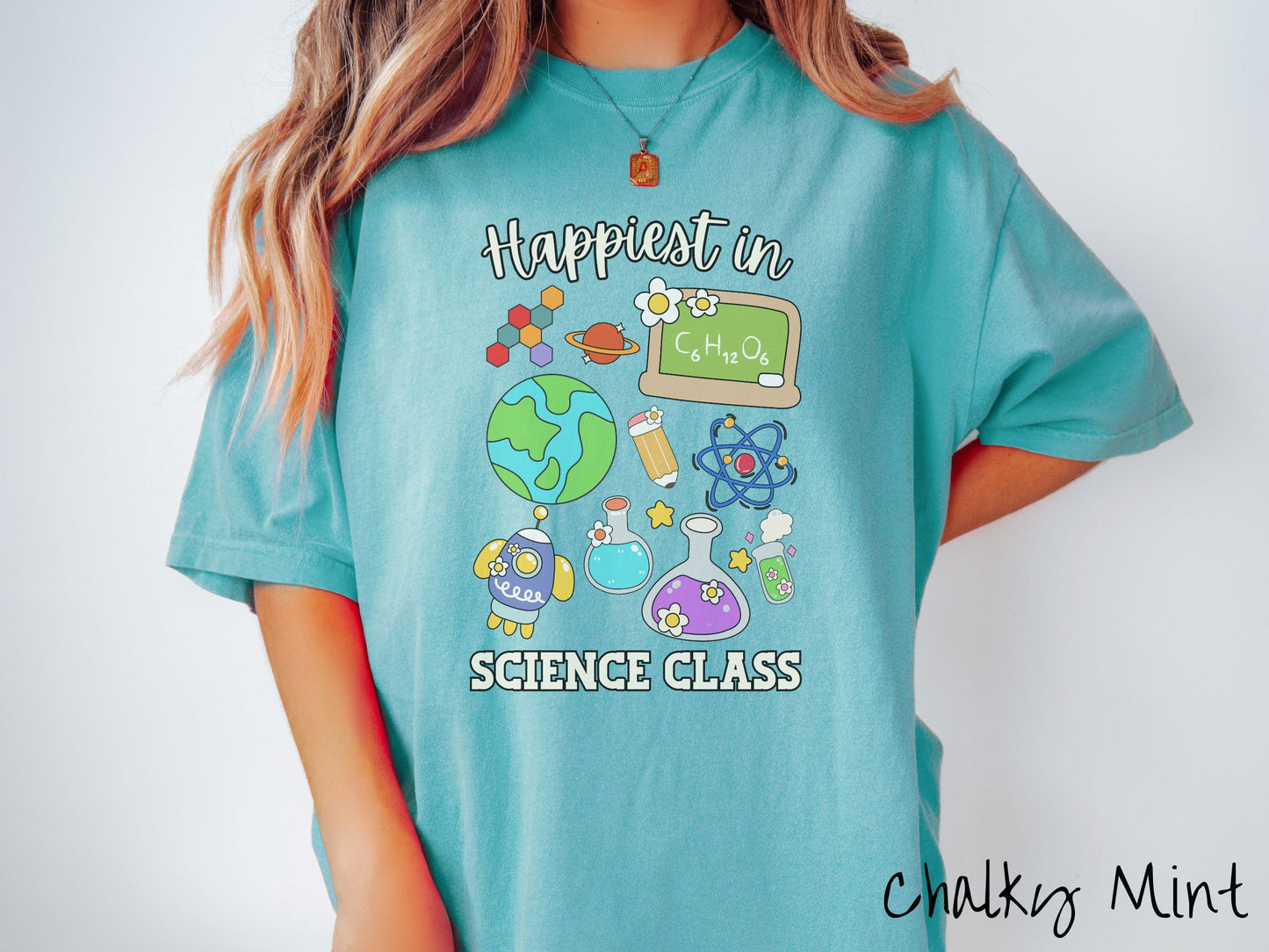 A woman wearing a cute, vintage chalky mint colored Comfort Colors T-shirt with the text Happiest in Science Class. In between the text are a globe, chemistry, rocket ship, chalkboard, science test tubes, a pencil, DNA molecules, and Saturn.