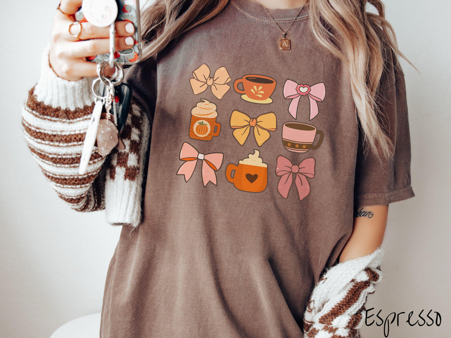 A woman wearing a cute, vintage espresso colored Comfort Colors T-shirt with a grid of pictures of an orange bow, an orange tea cup, a pink bow, an orange pumpkin-spiced latte, a yellow bow, a pink coffee cup, a pink bow, and an orange coffee drink.