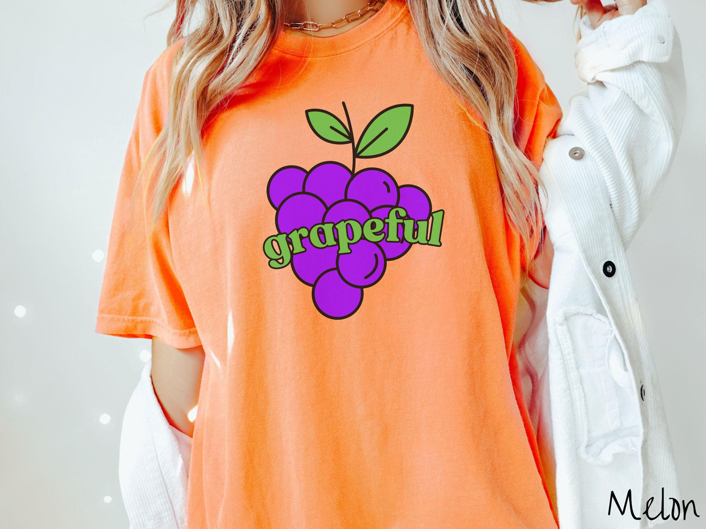 A woman wearing a cute, vintage melon colored Comfort Colors T-shirt with the text grapeful in green font across the front. Behind grapeful is a bunch of juicy, purple grapes with a green stem.