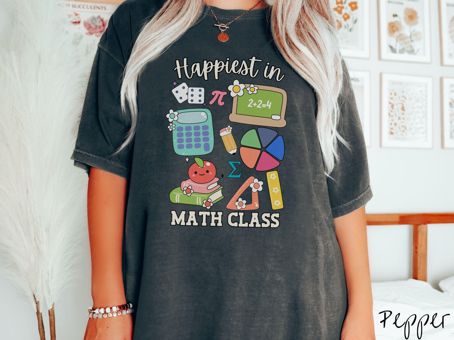 A woman wearing a cute, vintage pepper colored Comfort Colors T-shirt with the text Happiest in Math Class. In between the text are the pi symbol, a chalkboard, calculator, pencil, books, protractor, ruler, dice, an apple, and flowers.