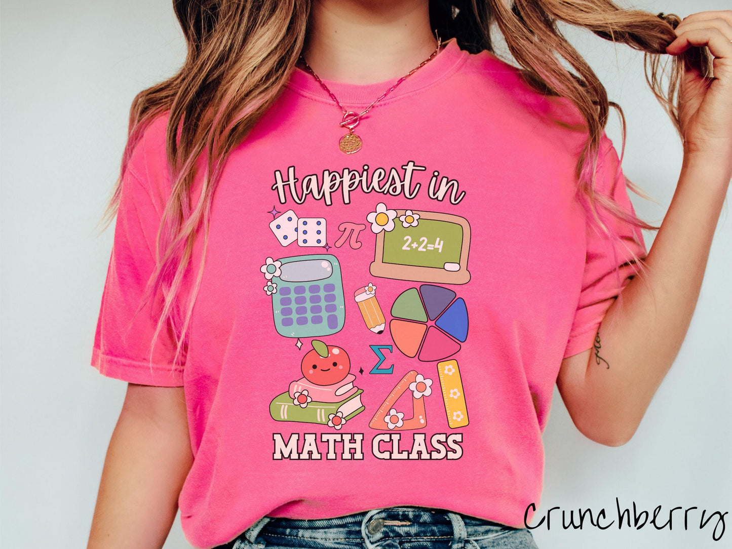 A woman wearing a cute, vintage crunchberry colored Comfort Colors T-shirt with the text Happiest in Math Class. In between the text are the pi symbol, a chalkboard, calculator, pencil, books, protractor, ruler, dice, an apple, and flowers.