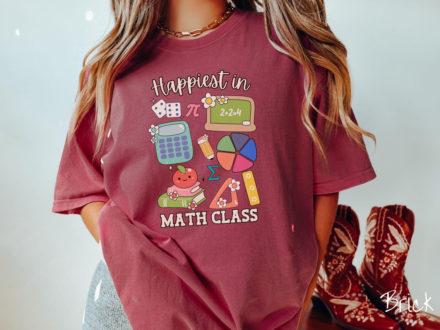 A woman wearing a cute, vintage brick colored Comfort Colors T-shirt with the text Happiest in Math Class. In between the text are the pi symbol, a chalkboard, calculator, pencil, books, protractor, ruler, dice, an apple, and flowers.