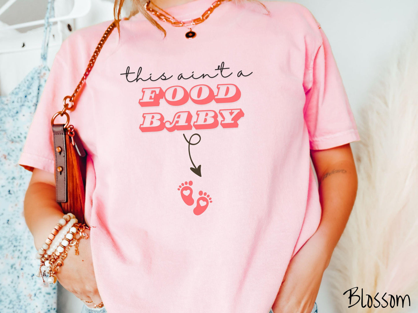 A woman wearing a cute, vintage blossom colored Comfort Colors T-shirt with the text this aint a food baby in black and red font. There is a black arrow pointing down to a pair of red baby footprints with hearts in them.