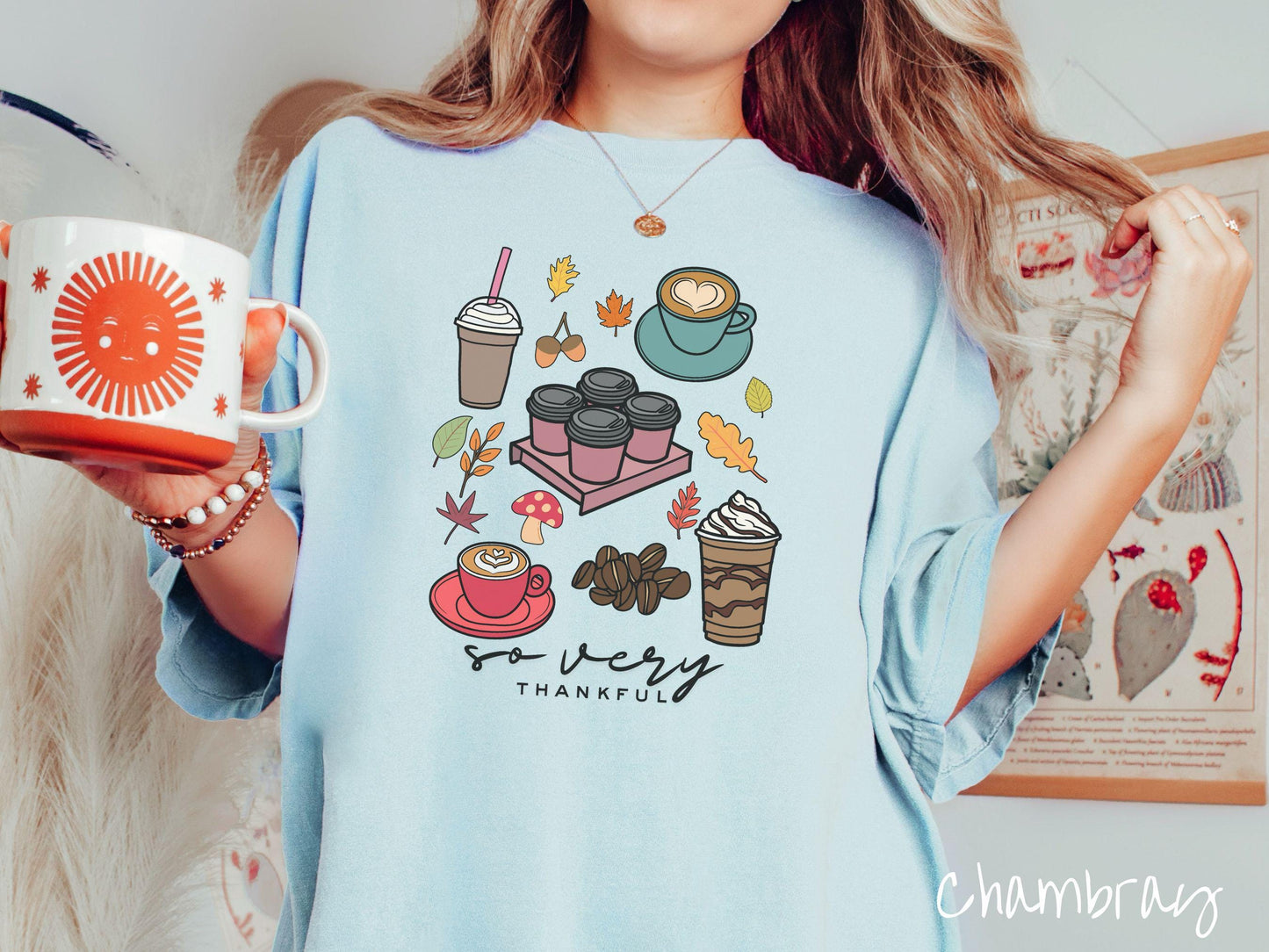 A woman wearing a cute, vintage chambray colored Comfort Colors T-shirt with the text so very thankful. Above are an iced coffee, four to-go cup coffees, a latte, a frappucino, coffee beans, mushrooms, and fall leaves scattered around.