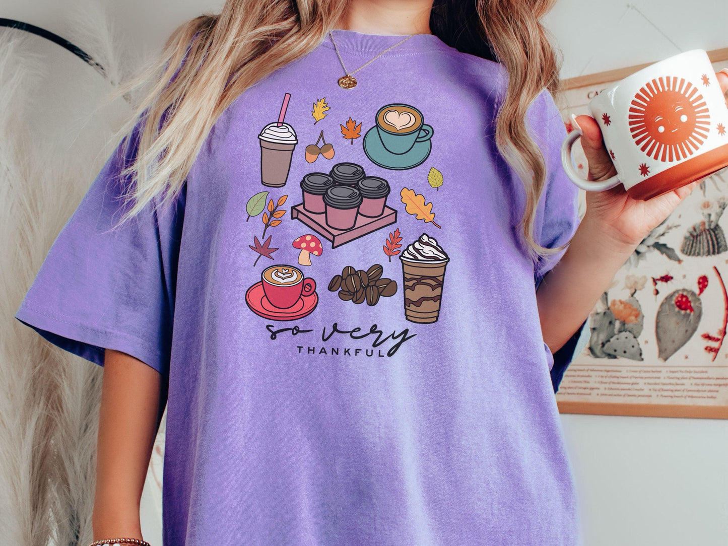 A woman wearing a cute, vintage violet colored Comfort Colors T-shirt with the text so very thankful. Above are an iced coffee, four to-go cup coffees, a latte, a frappucino, coffee beans, mushrooms, and fall leaves scattered around.