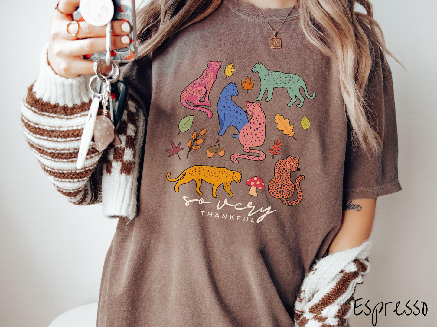 A woman wearing a cute, vintage espresso colored Comfort Colors T-shirt with the text so very thankful in black font. Above are colorful, spotted leopards in seated and walking positions, as well as leaves, acorns, and mushrooms scattered about.