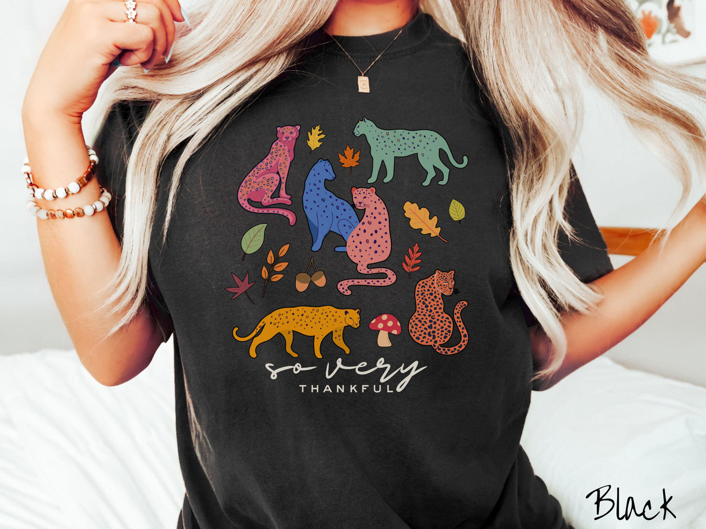 A woman wearing a cute, vintage black colored Comfort Colors T-shirt with the text so very thankful in black font. Above are colorful, spotted leopards in seated and walking positions, as well as leaves, acorns, and mushrooms scattered about.