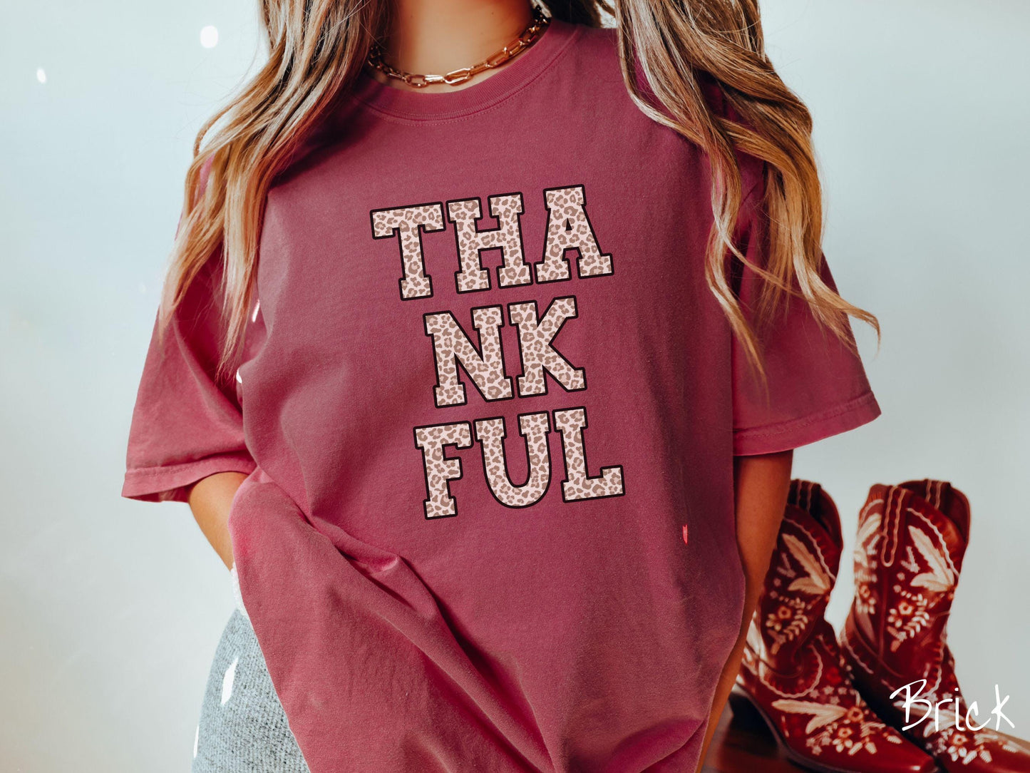 A woman wearing a cute, vintage brick colored Comfort Colors T-shirt with the text THANKFUL in capital letters stacked in three rows across the front. The letters are in ivory, light brown, and dark brown colored leopard print font.