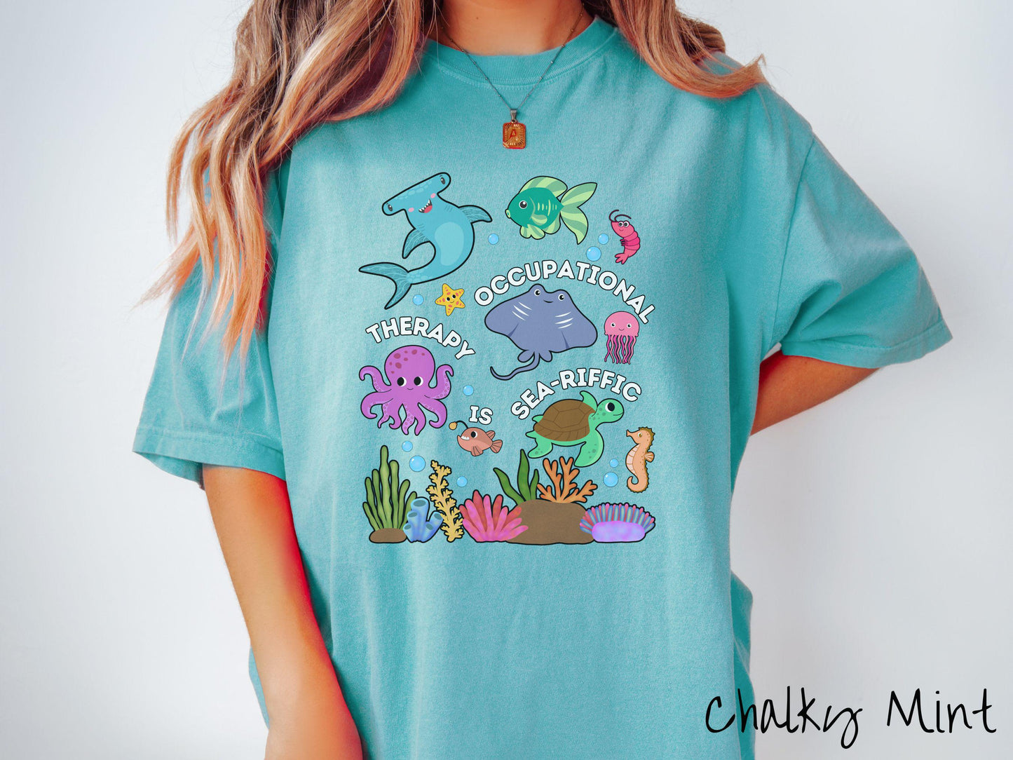 A woman wearing a cute, vintage chalky mint colored Comfort Colors T-shirt with text Occupational Therapy is Sea-Rific. There is a shark, green fish, octopus, stingray, jellyfish, turtle, and seahorse swimming together above a colorful coral reef.